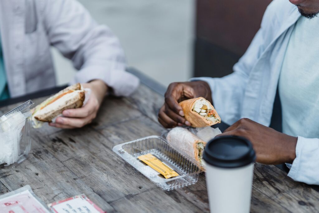 Two friends enjoying wraps and sandwiches at a rustic outdoor wooden table, sharing a casual lunch.